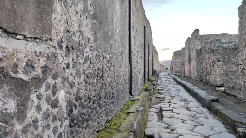 Slow pan right of a narrow stone street in Pompeii, Italy at golden hour, showing aged walls and cobblestones from the historic Roman city.
