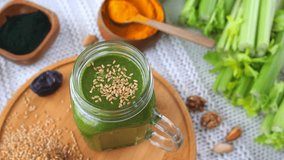 Healthy green smoothie in a glass jar rotating on a wooden plate, surrounded by ingredients like celery, spirulina, turmeric, nuts, and sesame seeds for a nutritious vegan breakfast - Powered by Shutterstock - Get 15% off with code: PIKWIZARD15