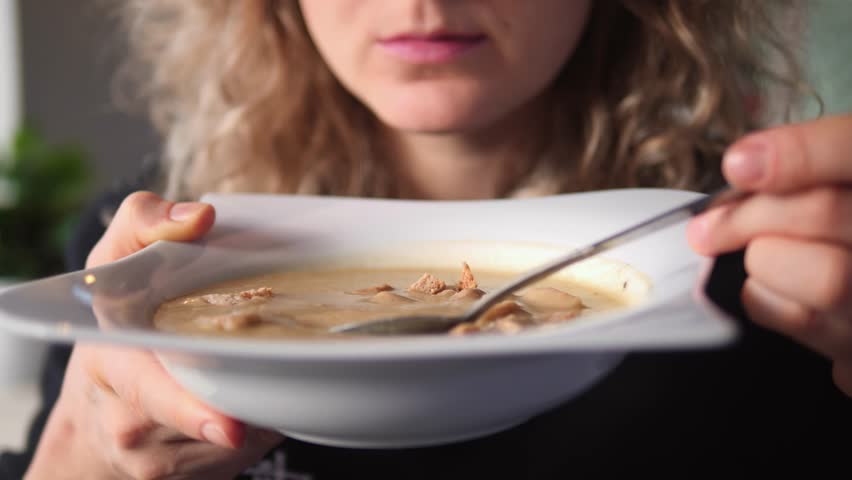 Close up of a young woman's mouth eating hot, creamy mushroom soup with meat from a white bowl. The curly haired person is holding a spoon and enjoying a homemade meal for lunch or dinner