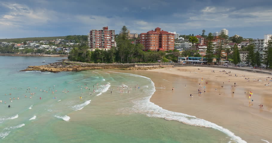 Sydney, Manly Beach coastline with turquoise water, people relax, swim and sunbath on golden sand, beachfront buildings and esplanade under bright blue sky, ideal seaside scene. Aerial view flight