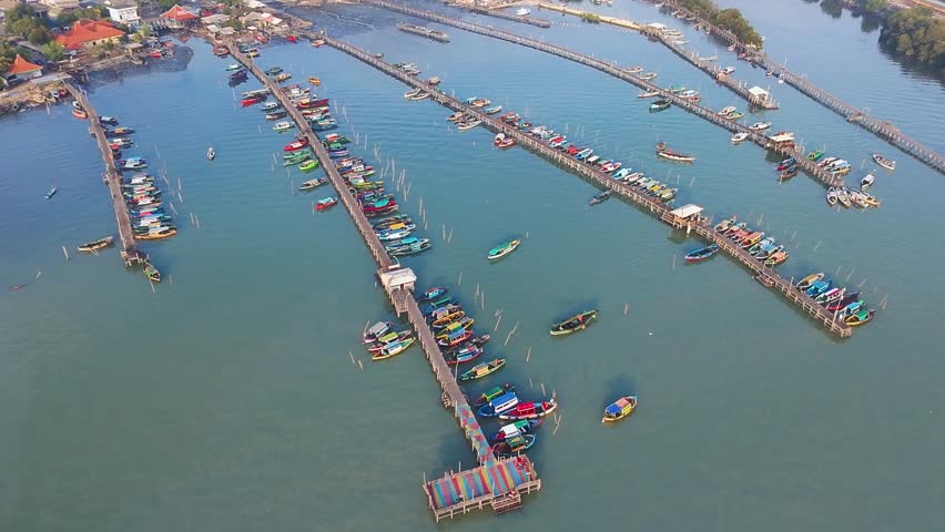Aerial view of vibrant fishing boats docked at long wooden piers in a harbor near Lumpur, Gresik, East Java, with a dense coastal town skyline.