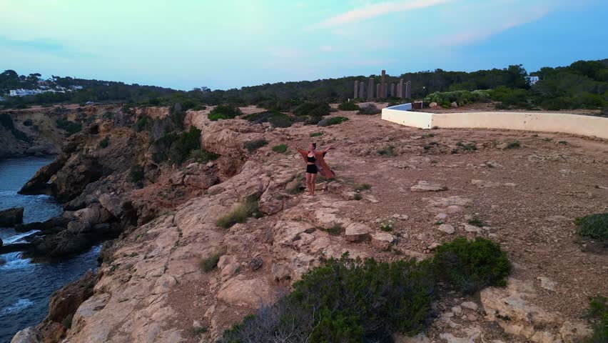 dancing Woman expressing freedom and movement by the sea at a beautiful travel destination Cala Llentia Ibiza. Nice aerial view flight static tripod hovering drone