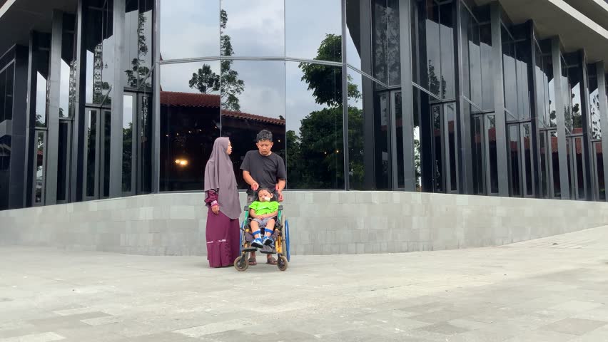 loving family strolls outdoors, with parents guiding their child in a wheelchair past a contemporary building featuring striking, reflective glass panels.