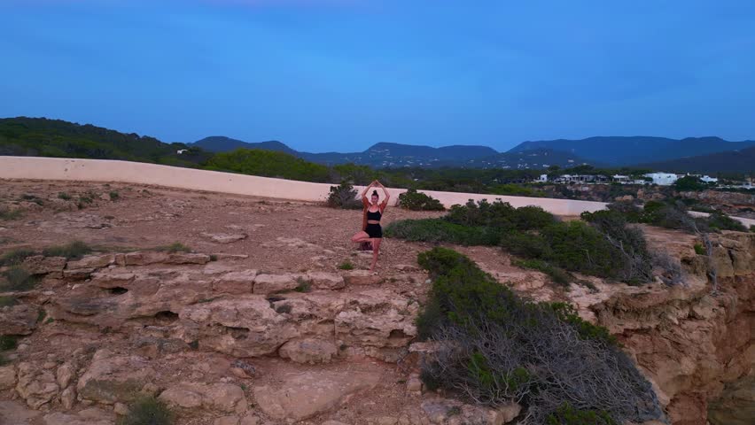 Woman performing a yoga tree pose on a rocky cliff during a serene sunset in Cala Llentia Ibiza. Fantastic aerial view flight circle drone footage