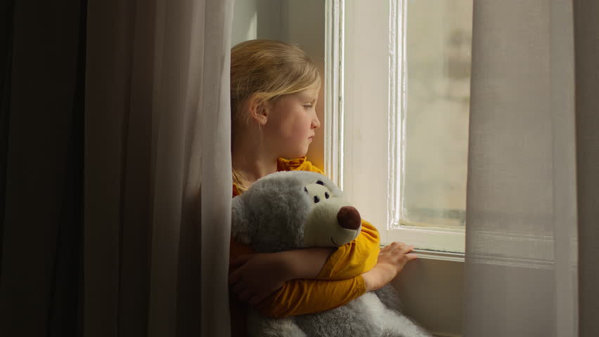 Frustrated lonely Caucasian girl child kid orphan daughter looking out window holding teddy bear bored victim innocence depressed alone waiting for family adoption custody ill sick quarantine at home