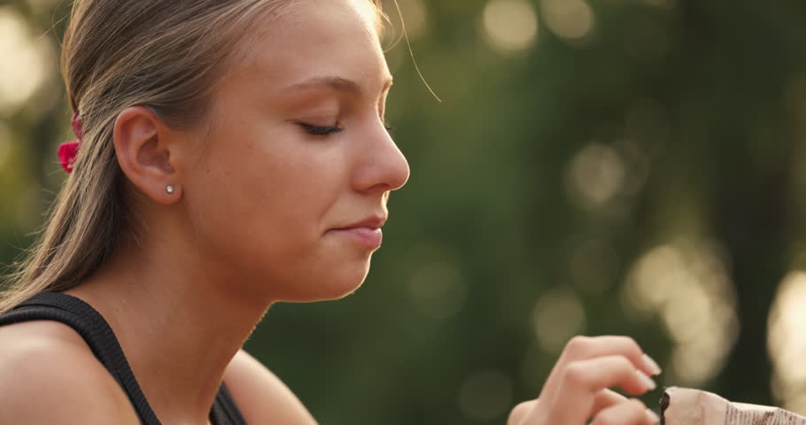 A girl takes a bite of a crunchy snack while relaxing outside. She sits in a natural setting, with sunlight filtering through trees, creating a peaceful atmosphere.