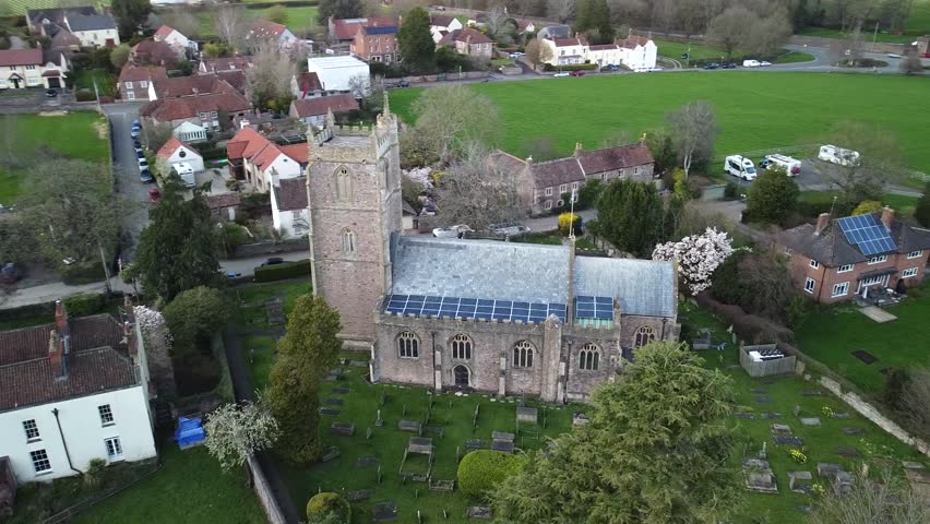 Historic village church aerial view in rural England. Scenic aerial drone footage of a stone church surrounded by a traditional English village and countryside. Churchyard with old gravestone.
