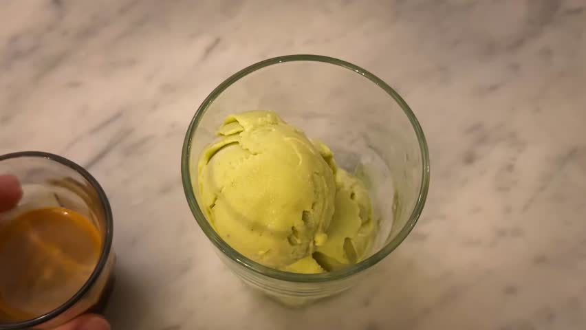 Close-up video of a hand pouring freshly brewed espresso over a scoop of green ice cream to make affogato. Creamy texture, warm coffee color, and marble background create a cozy café atmosphere.
