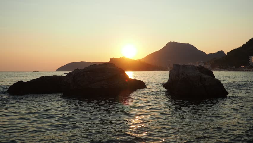 Golden sun sets on calm Adriatic waters in Sutomore, Montenegro. Large rocks dot the shoreline while silhouettes of mountains frame this picturesque Mediterranean landscape