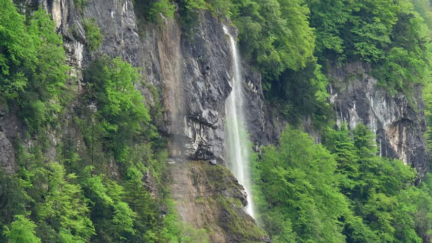 Waterfall cascades down rocky cliff in lush green landscape near mountain range