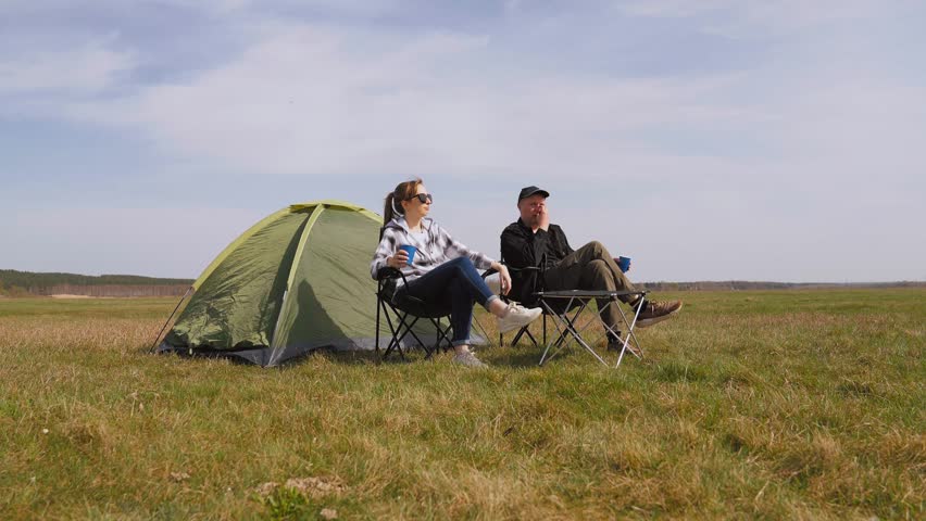 Happy couple sitting in camping chairs near their tent, drinking from cups and enjoying the beautiful spring scenery during a relaxing weekend getaway in the great outdoors