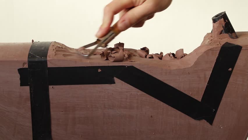An industrial designer shaving off pieces of automotive clay from a roughly shaped design model using a sculpting tool.