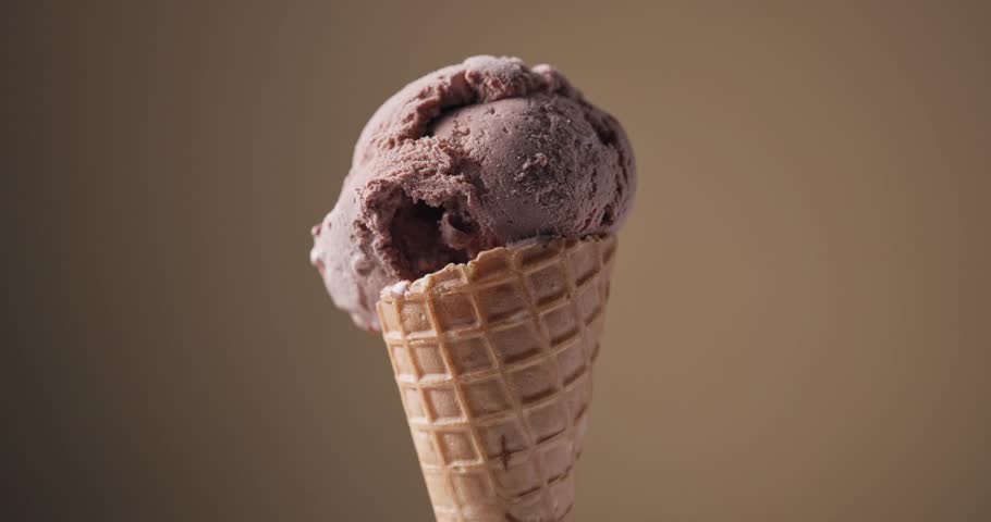 Melted chocolate sauce flowing over a scoop of chocolate ice cream in a waffle cone, set against a brown background