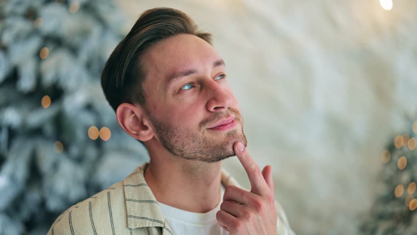 Thoughtful young man contemplating, finger on chin. Reflecting with hopeful expression, pleasant thoughts fill mind. Upward gaze and festive background evoke dreaming mood. Deep thought, peaceful.