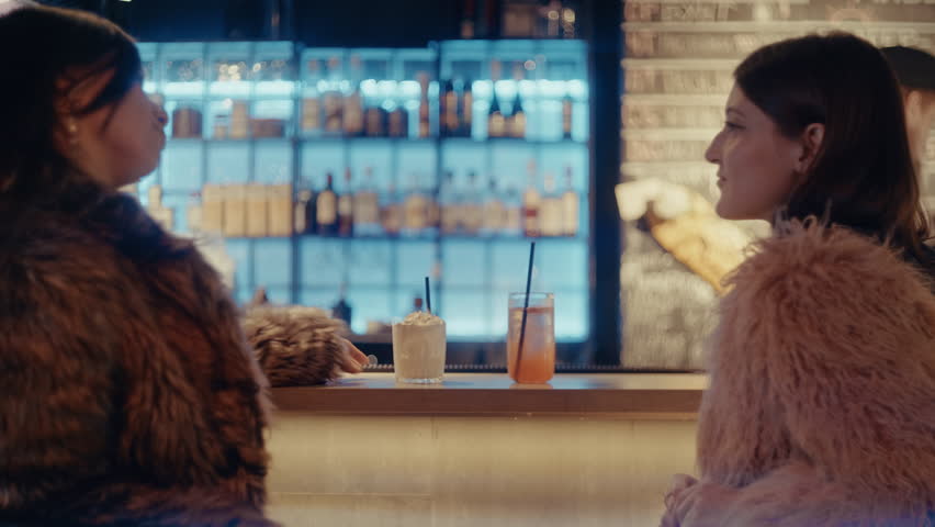 A close-up shot of two young women clinking cocktail glasses and making a toast at a modern bar counter during the evening.