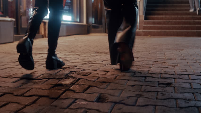 Two fashionable women in stylish boots walk along a cobbled street at night, heading toward a bar.
