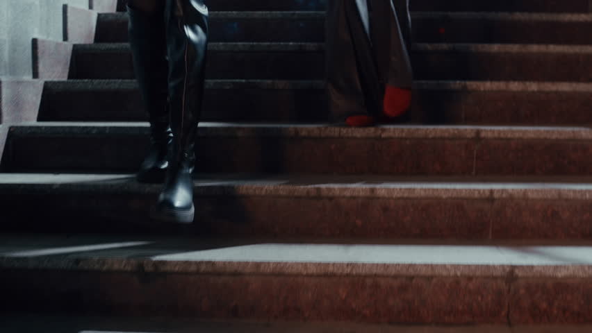 Two fashionable young women in faux fur coats and stylish boots walk down the stairs illuminated by city lights and enter a bar for a weekend night out.