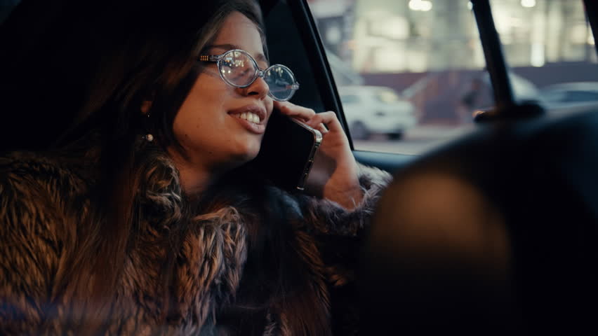 Fashionable female passenger rides through the city at night, chatting on her smartphone with a smile.
