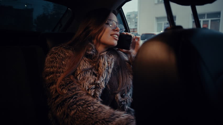 A glamorous young woman wearing a fur coat smiles while talking on her smartphone in the backseat of a taxi at night.