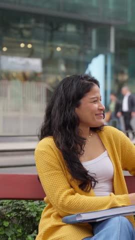 Two cheerful young female students sitting on a bench outdoors, working together on a laptop and talking after their classes
