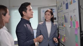 Three people are standing in front of a white board with various graphs and numbers. One of the men is pointing at the board, while the other two women look on - Powered by Shutterstock - Get 15% off with code: PIKWIZARD15