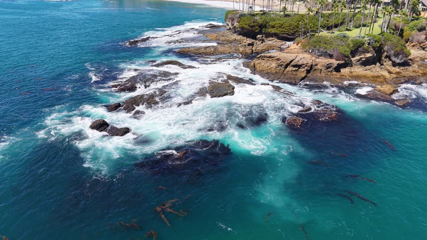 Kelp forests swirl beneath crashing waves along the rocky shoreline of Laguna Beach, captured from an overhead drone perspective