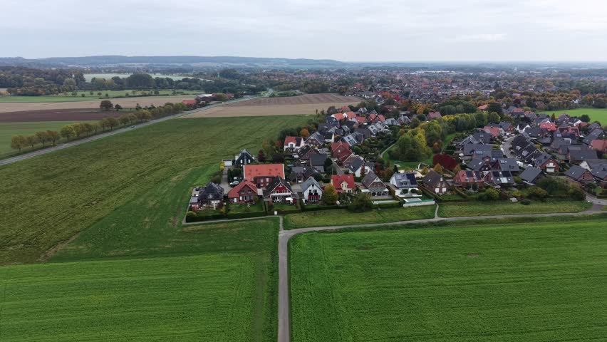 European rural suburb neighborhood with green farmland fields and single family houses. Aerial lateral wide shot. Cloudy fall day in October. Germany, Europe. Panorama view. Quiet scene.