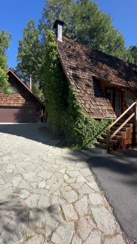 Rustic A-Frame Cabin Exterior with Cedar Shake Siding, Stone Driveway, Wooden Porch Stairs, Ivy-Covered Wall, and Surrounded by Tall Pine Trees Under Bright Blue Sky in Mountain Forest Setting