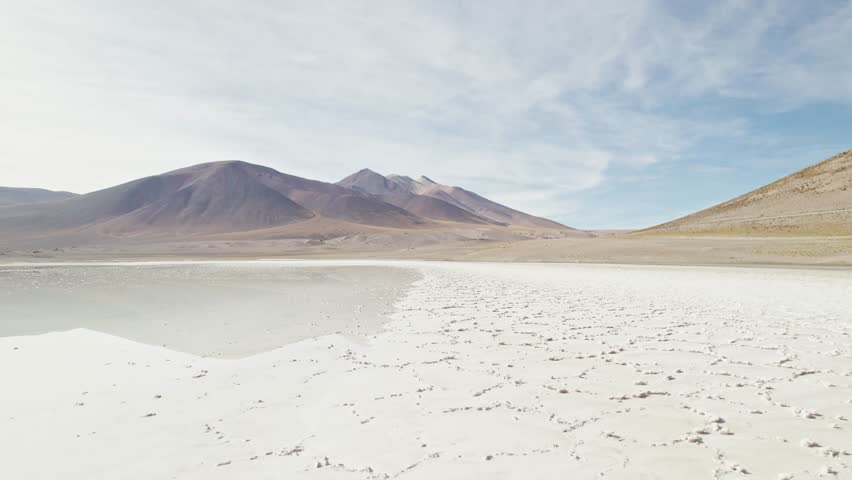 Aerial view of a salt flat and surrounding mountains in the Atacama Desert, Chile. White mineral crusts contrast with volcanic peaks and a clear blue sky