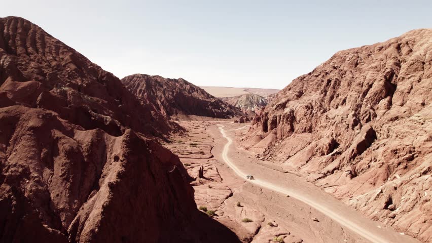 Drone following a car through the red canyons of the Atacama Desert, Chile. Rugged sandstone cliffs, winding dirt road, and bright daylight create a cinematic desert scene