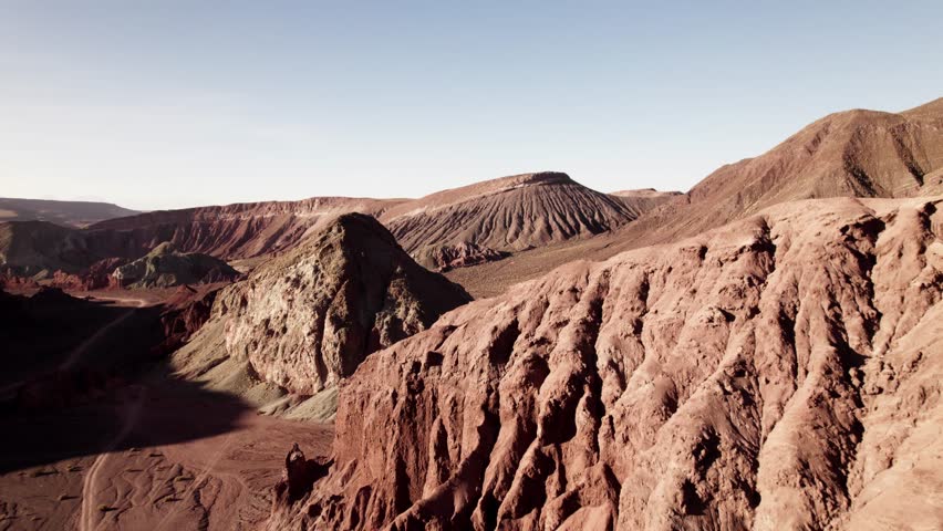 Aerial view of layered volcanic hills in the Atacama Desert, Chile. The eroded red and green formations, dry terrain, and clear sky create a surreal and ancient Martian landscape