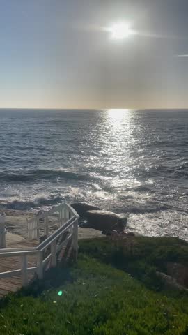 Rocky Coastline with Ocean Waves Sparkling Under the Sun and a Wooden Staircase Leading Toward the Water Surrounded by Green Coastal Plants, White Wooden Fence and Clear Blue Sky