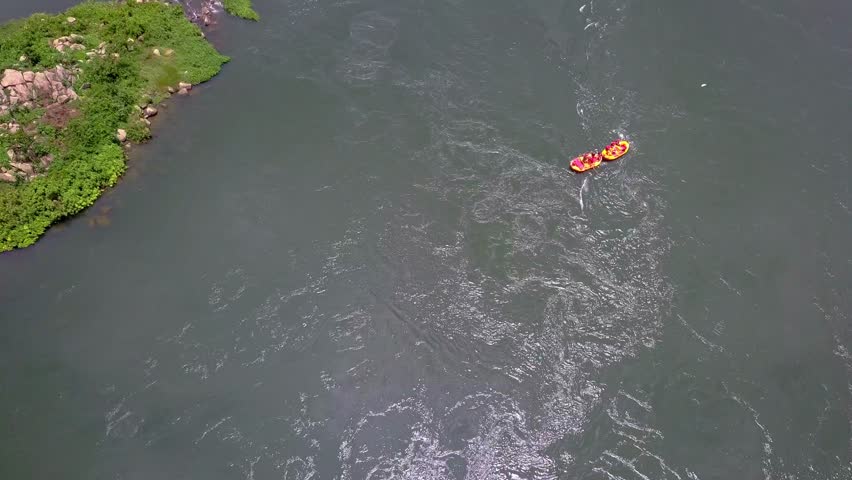 Top‑down aerial view of two inflatable rafts navigating the River Nile in Uganda, with rafters paddling downstream past green riverbanks and rocky edges.