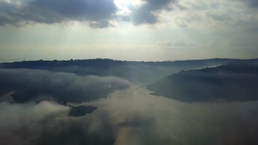 Aerial wide drone view of Lake Bunyonyi in southwestern Uganda, with mist and low lying clouds drifting over the water and terraced hills, illuminated by soft sunlight.