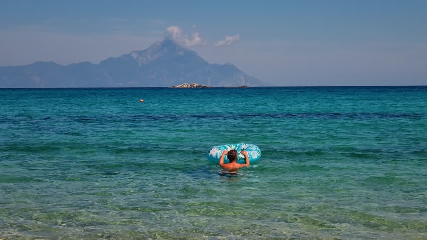 Man floating with an inflatable ring in the crystal-clear turquoise waters of Kriaritsi, Greece, enjoying a sunny Mediterranean summer holiday