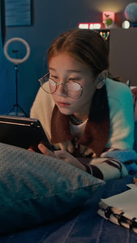 Vertical shot of focused multicultural gen Alpha girl in eyeglasses using digital tablet when lying in bed and doing homework at night