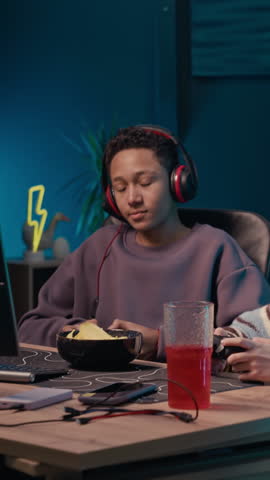 Vertical shot of gen Alpha African American boy sitting next to his peer at table, wearing wireless headphones while video gaming on computer late at night