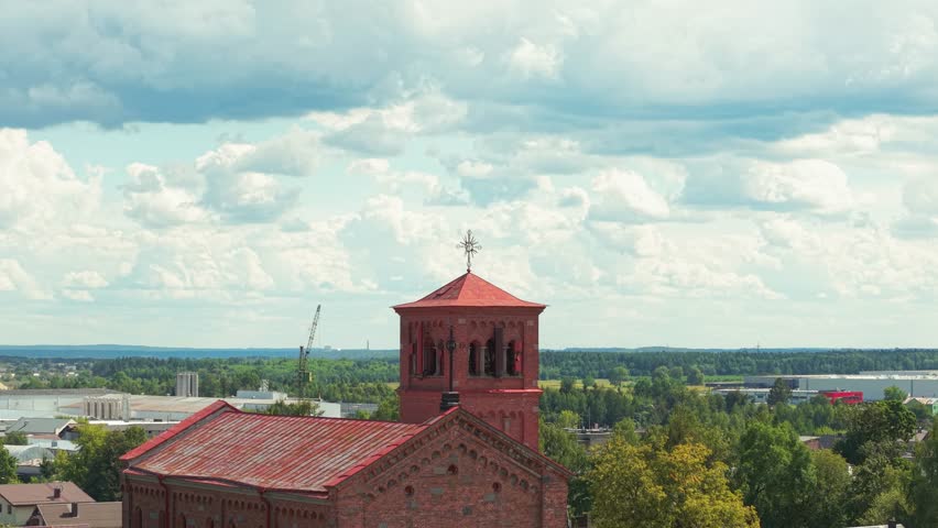 Aerial view of Lentvaris Church in Lithuania with its red brick architecture and bell tower, surrounded by lush greenery and a distant cityscape under a partly cloudy sky.
