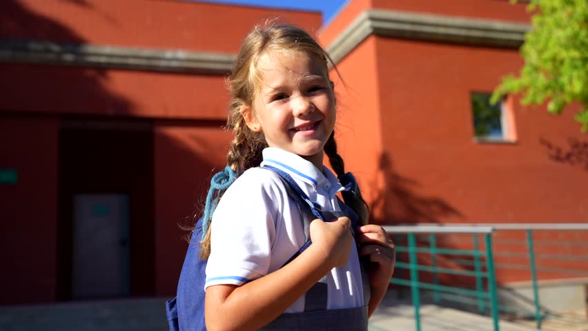 Beautiful elementary school girl with pigtails and a backpack smiling and waving goodbye to her mother before entering the school building on a sunny day, ready for a new day of learning