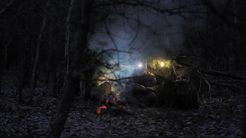 Night camp with survival in the forest under a pine canopy. A man with a headlamp drinks from a metal cup by the campfire in a survival camp in the forest, lit by a lantern at night.