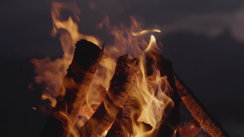 Bright campfire burning in the foreground with a mountain range and lake in the background at night. Peaceful outdoor scene symbolizing warmth, adventure, and solitude in nature.