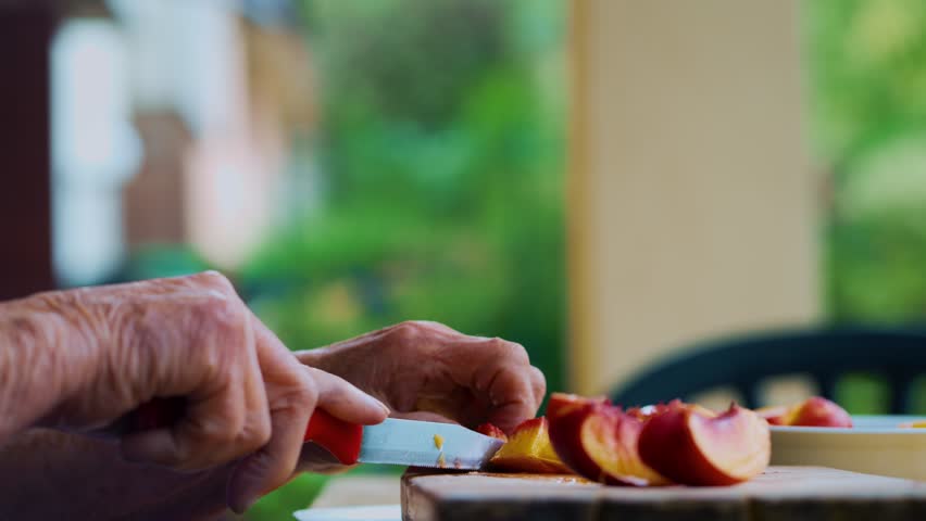Senior woman slices a fresh peach with a knife on a rustic table outdoors surrounded by greenery