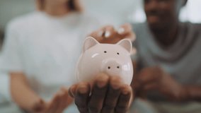 Closeup of funny ceramic piggy bank in hand of black man, woman putting coins. People save money for buying house and dreams realizing, family budget and finance, happy multiracial married couple - Powered by Shutterstock - Get 15% off with code: PIKWIZARD15