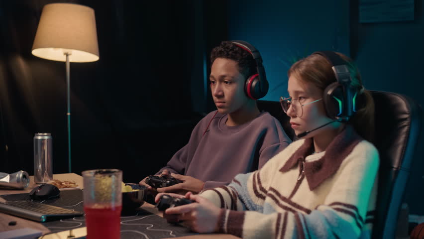 Medium shot of two diverse gen Alpha gamers in wireless headphones sitting at computer table when playing console and eating snacks late at night