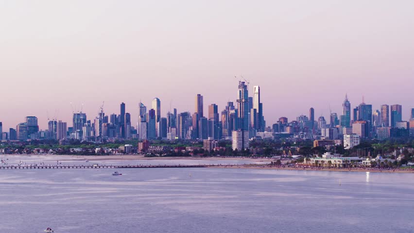 A beautiful aerial shot flying sideways over Port Phillip Bay at dusk, revealing the St Kilda pier and beach area with the Melbourne CBD skyline illuminated in the distance.