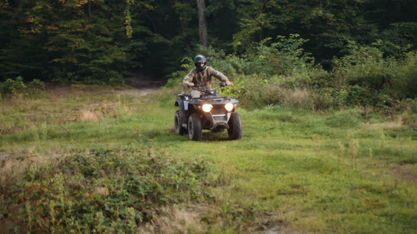 Dense forest, All-terrain rider, Trail navigation. Camouflaged ATV operator with helmet navigates dirt path in dense forest, using headlights to guide way.