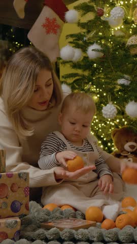 Vertical Screen: A mother and her young child sit on a cozy blanket, playing with bright oranges next to a beautifully decorated Christmas tree, creating joyful holiday memories.