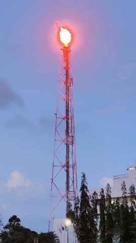 An industrial scene featuring a tall oil and gas refinery tower with a bright flare burning at its peak. This image symbolizes the complex process of extracting and managing energy resources.