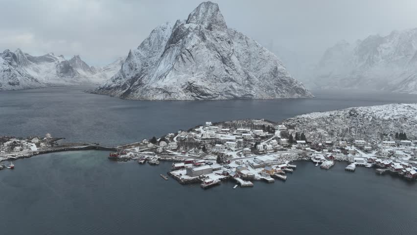 Aerial view of snow-covered houses nestled on a hillside, with majestic mountain ranges and tranquil water, creating a serene winter scene, Norway.