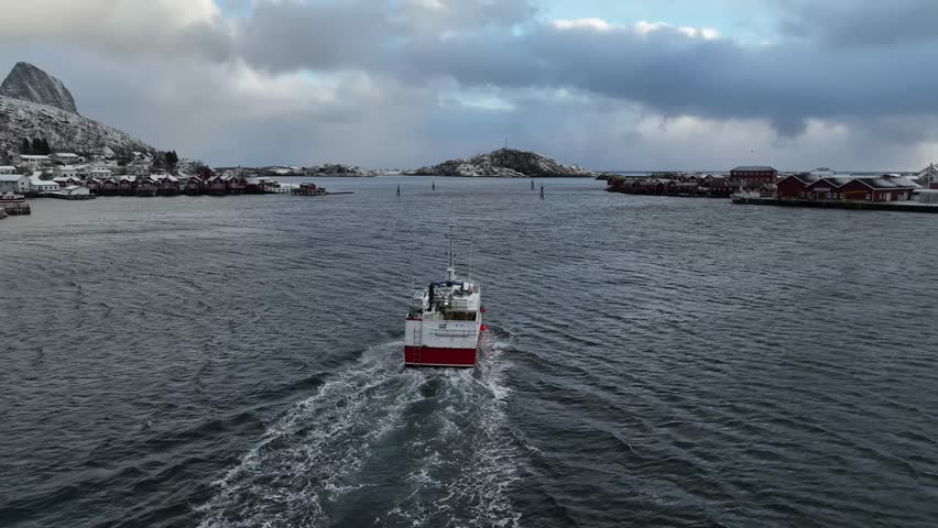 Aerial view of a red and white fishing boat leaving Reine, amidst dark waters contrasting with the snowy mountains, Reine, Norway.